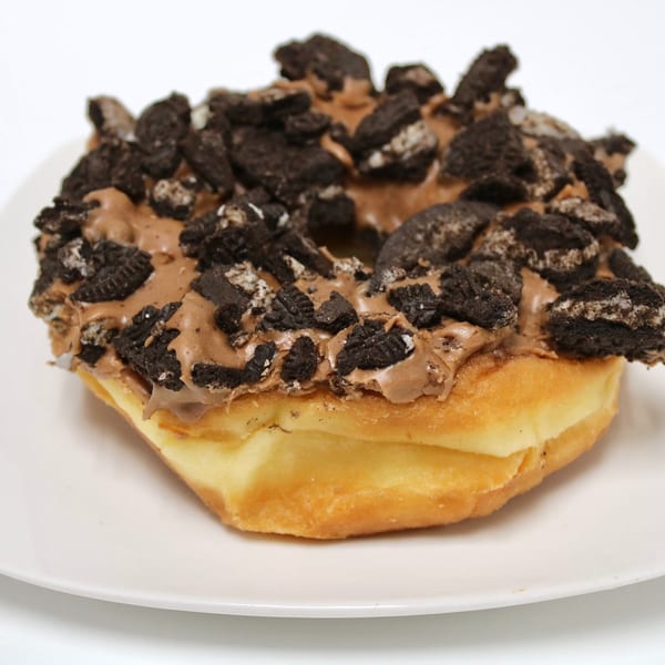 A golden-brown donut topped with chocolate frosting and crushed Oreo cookies on a white plate.