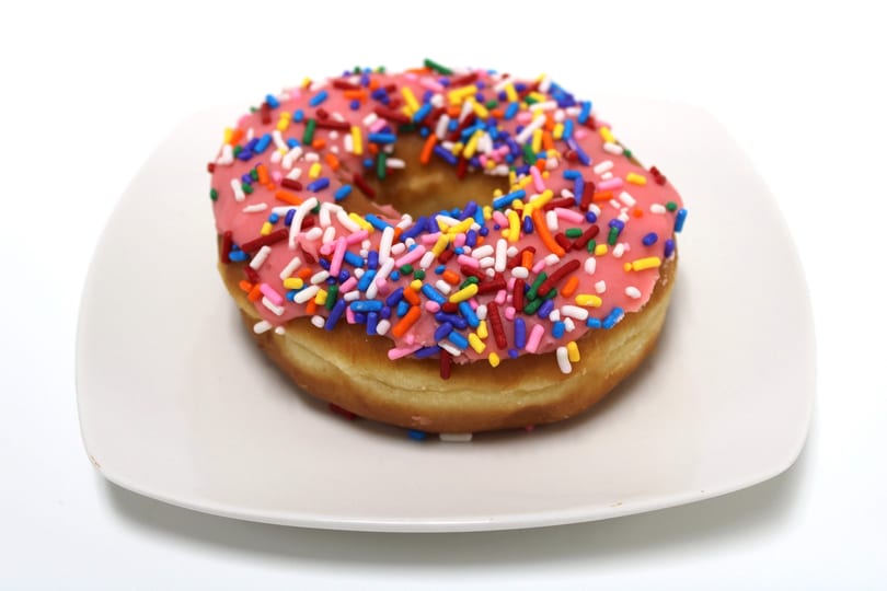 Pink frosted donut with colorful sprinkles on a white plate.