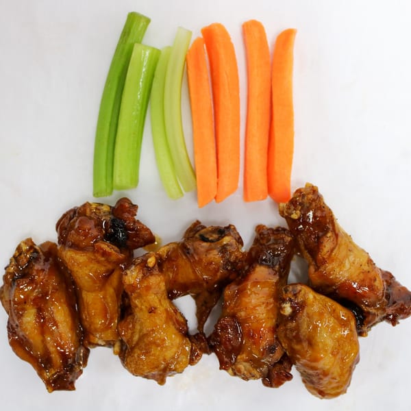 Glazed chicken wings with celery and carrot sticks arranged on a white surface.