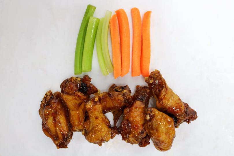 Glazed chicken wings with celery and carrot sticks arranged on a white surface.