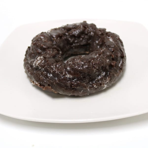 A dark chocolate old-fashioned donut with a shiny glaze sits on a white square plate.