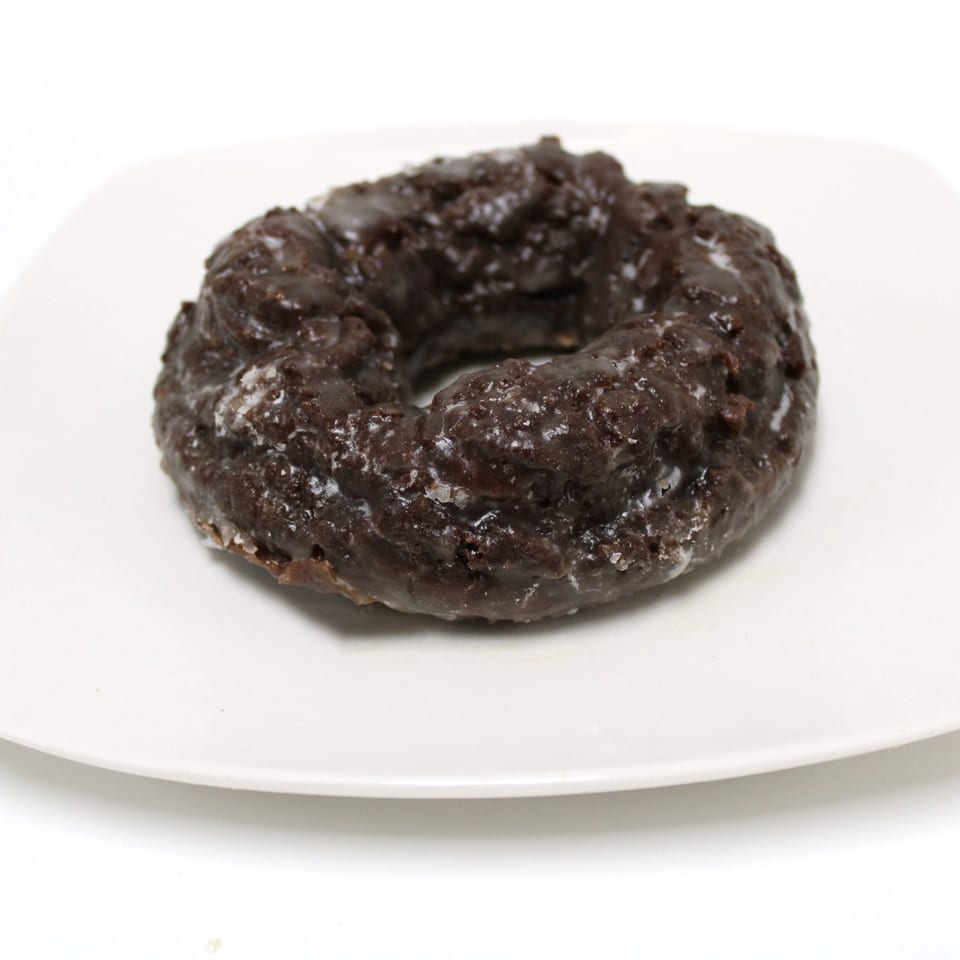 A dark chocolate old-fashioned donut with a shiny glaze sits on a white square plate.