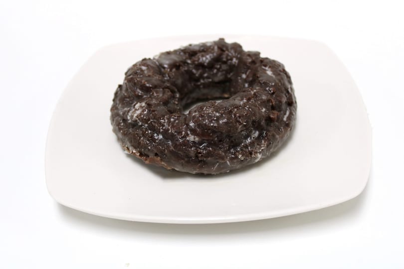A dark chocolate old-fashioned donut with a shiny glaze sits on a white square plate.