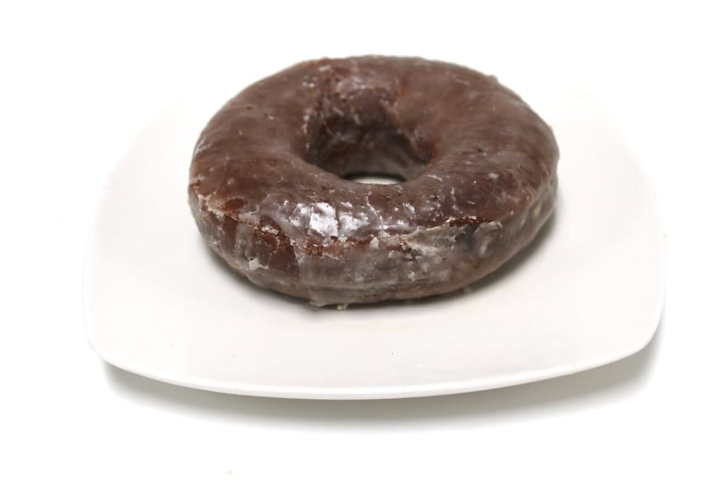 A single chocolate glazed donut sits on a white square plate against a white background.