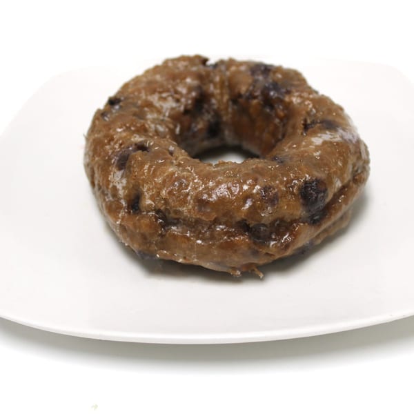 A glazed blueberry donut sits on a white square plate against a white background.