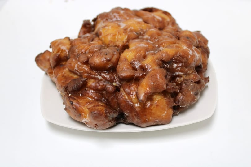 A large, glazed apple fritter with a golden-brown, craggy surface sits on a white plate.