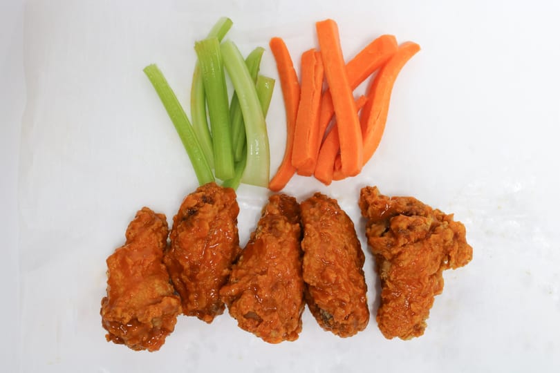 Five buffalo chicken wings with celery and carrot sticks on a white surface.